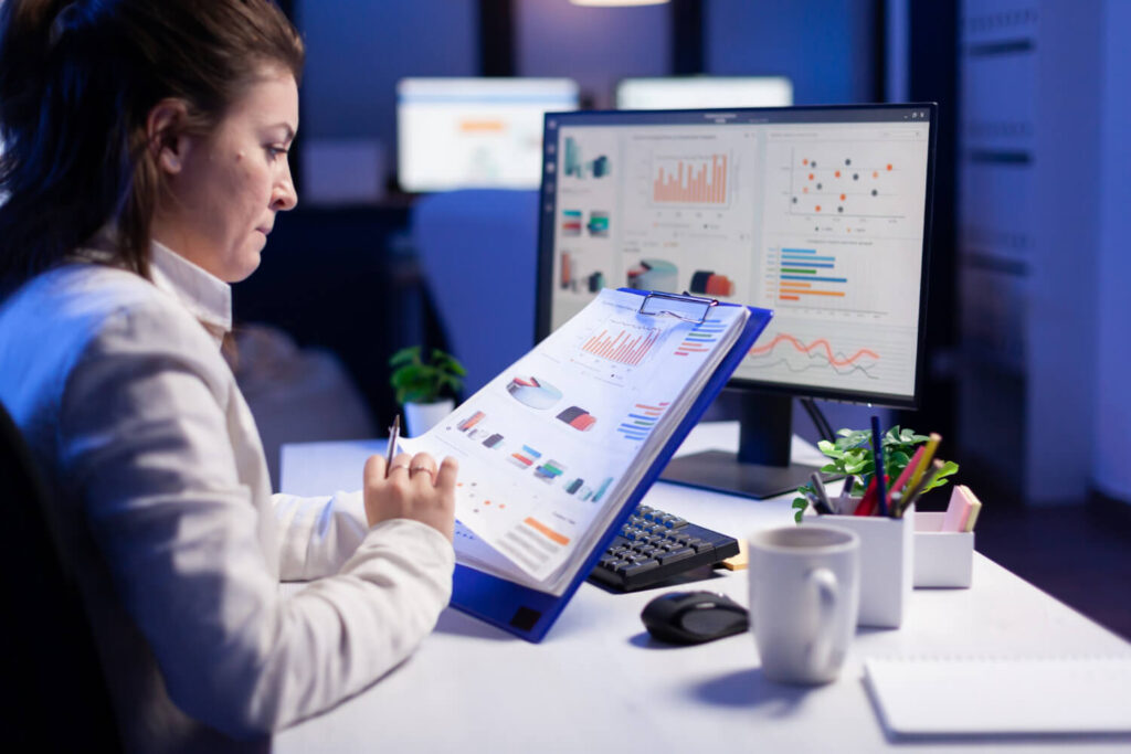 Freelancer woman comparing graphics from clipboard with grafics from computer at business office